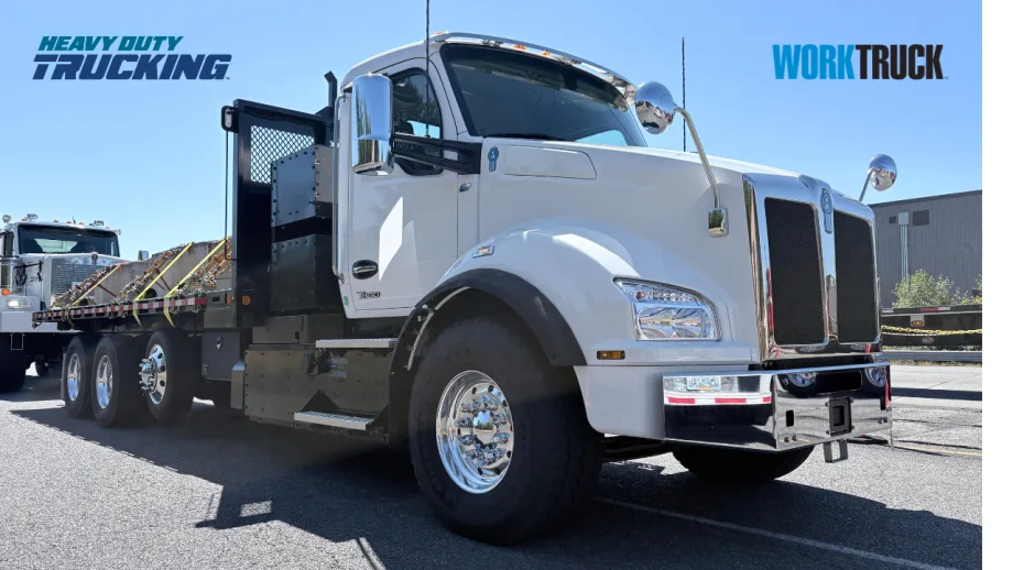 White Kenworth T880 heavy-duty flatbed truck displayed outdoors, featuring chrome accents and rugged tires, with another work truck visible in the background.