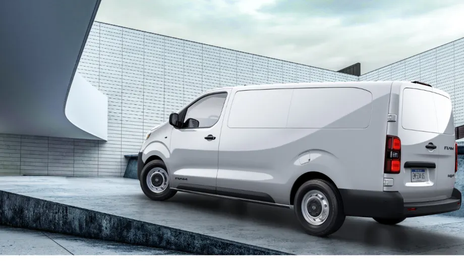 White Ram ProMaster City cargo van shown from the rear three-quarter view outside a modern industrial building, highlighting its windowless cargo area, rear doors, and compact commercial design.