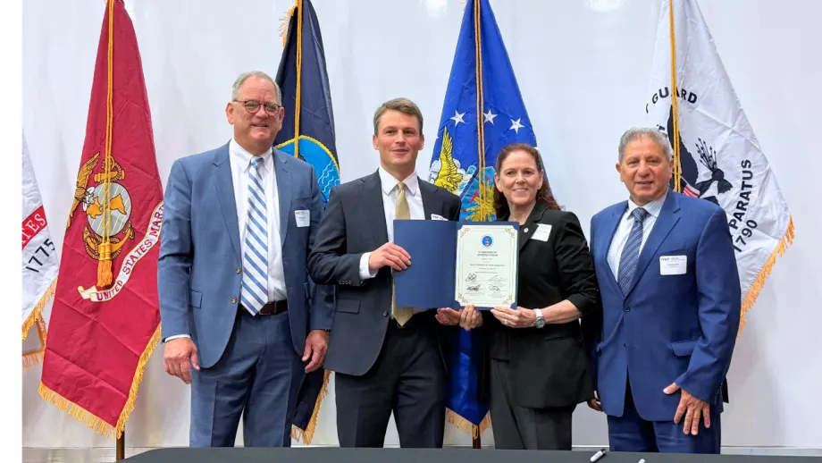 Four representatives pose with a signed certificate during an Isuzu and IVAP event, standing in front of U.S. military service flags.