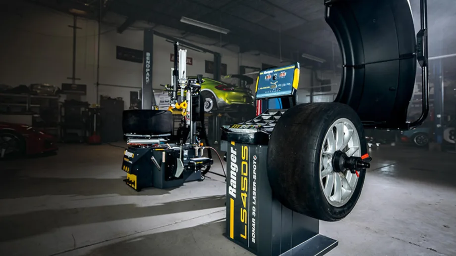 Ranger tire changer and wheel balancer equipment displayed in an automotive service shop, with a mounted performance tire and vehicles on lifts in the background.