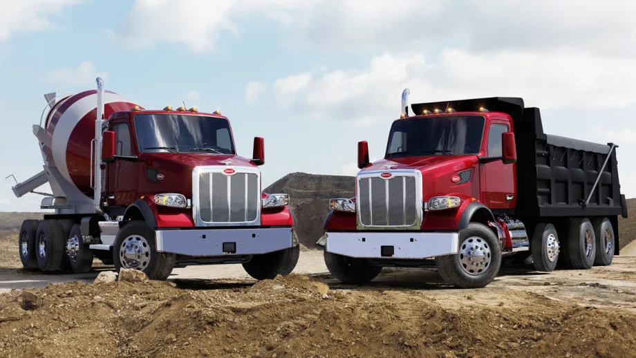 Two red Peterbilt vocational trucks, a concrete mixer and dump truck, parked at a construction site with dirt piles and a partly cloudy sky in the background.