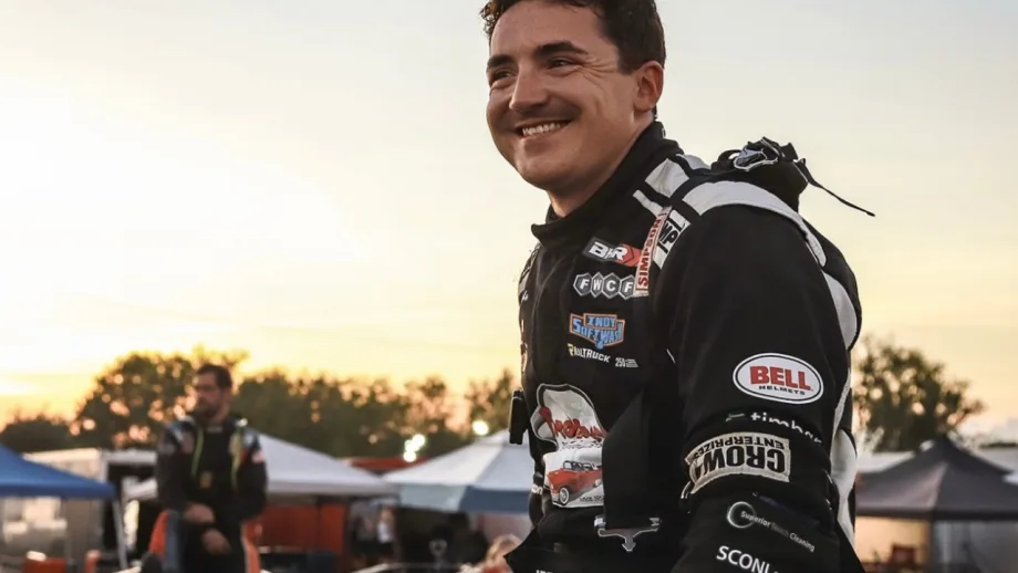 Isaac Johnson in a black-and-white firesuit smiles at the track at sunset with tents, equipment, and another person blurred in the background.