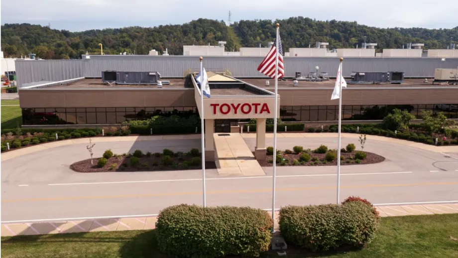 Aerial view of a Toyota manufacturing facility entrance, featuring the Toyota name above the doorway, three flagpoles, landscaped grounds, and the factory complex in the background.