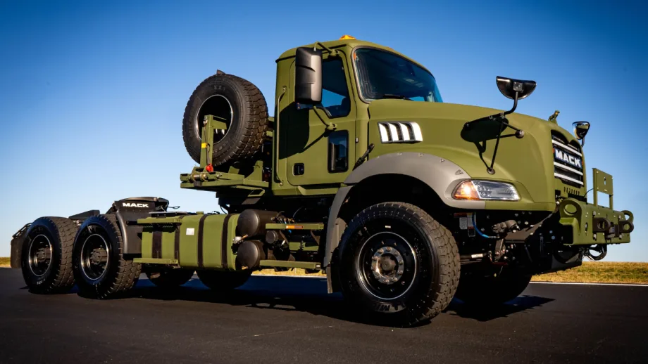 Olive-drab Mack Defense military tactical truck chassis with rugged tires, spare tire mounted behind the cab, heavy-duty front bumper, and three-axle configuration, photographed under a clear blue sky.
