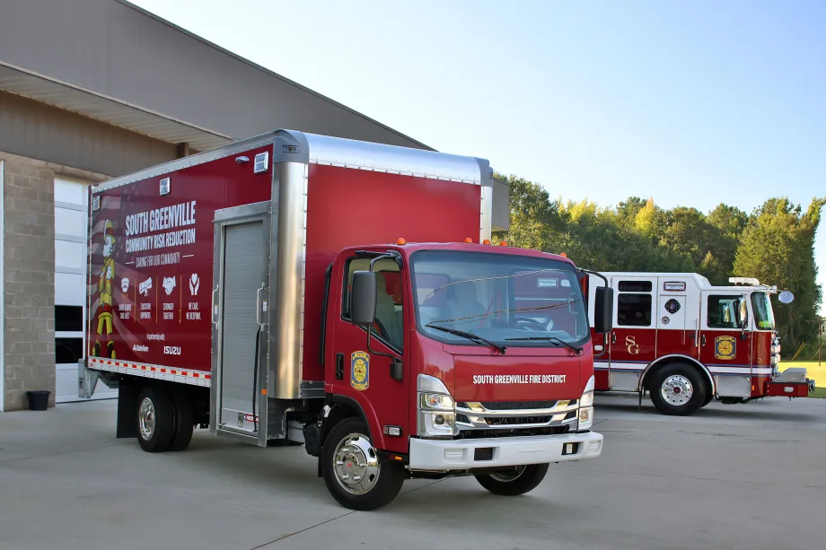 South Greenville Fire District red Isuzu box truck featuring Community Risk Reduction graphics, parked outside a fire station with a red-and-white fire engine in the background.