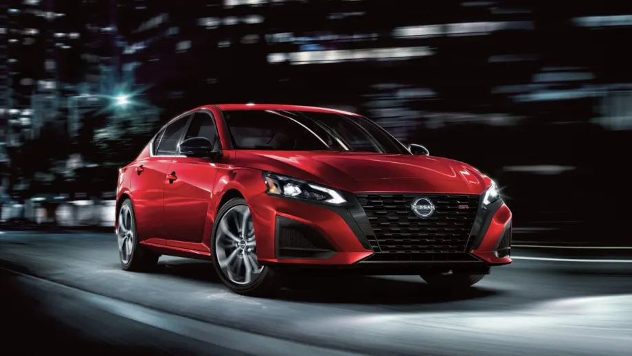 Red 2027 Nissan Altima SR sedan driving through a city at night, shown from the front three-quarter angle with illuminated headlights and dark alloy wheels.