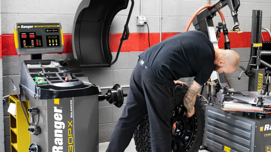 Automotive technician handling a truck tire beside a Ranger DST30PX professional wheel balancer and tire-changing equipment inside a vehicle service shop.