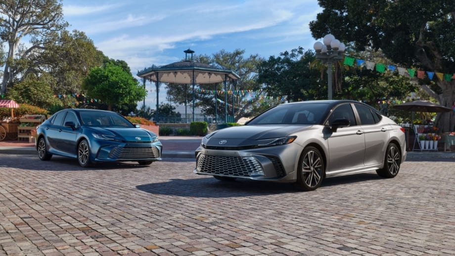 Two 2027 Toyota Camry sedans, one silver and one blue, parked on a cobblestone plaza with a gazebo, trees, and colorful decorations in the background.