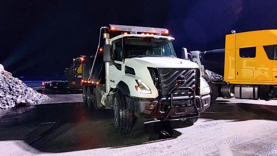 White Volvo VNX dump truck with illuminated roof lights, rugged tires, and front grille guard parked at a snowy nighttime worksite beside a yellow semi-truck and piles of rocks.