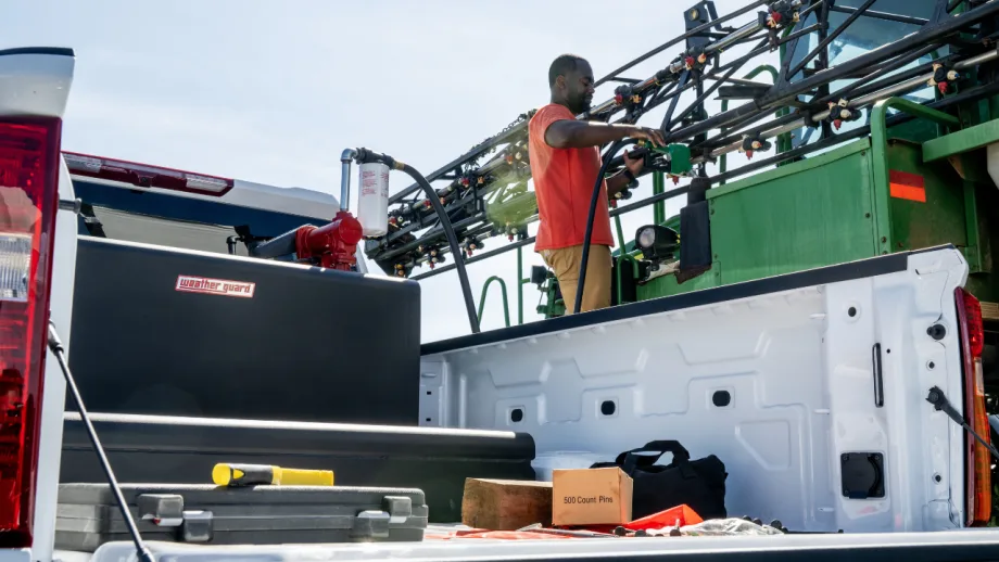 A worker refills agricultural spraying equipment beside a white pickup truck equipped with a Weather Guard transfer tank, with tools and supplies visible in the truck bed.