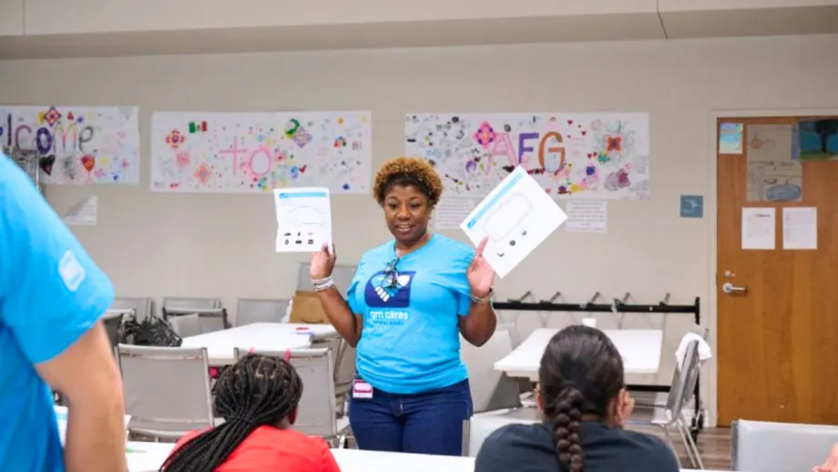 A woman wearing a blue GM Cares shirt holds up educational materials while speaking to a group of participants seated at tables during a community outreach or educational workshop.