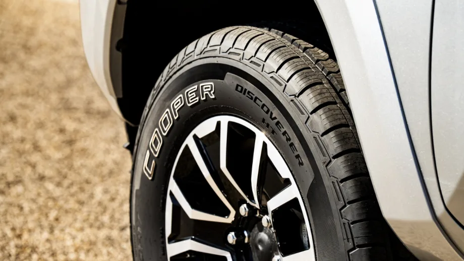 Close-up of a Cooper Discoverer HT tire mounted on a black-and-silver alloy wheel of a silver pickup truck, highlighting the tire’s tread pattern and Cooper branding on the sidewall.