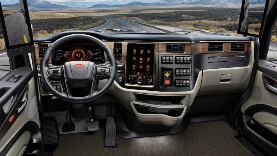 Interior view of a Peterbilt commercial truck cab featuring a digital instrument cluster, touchscreen infotainment system, steering wheel, dashboard controls, and digital side mirrors, with a winding highway and mountains visible through the windshield.