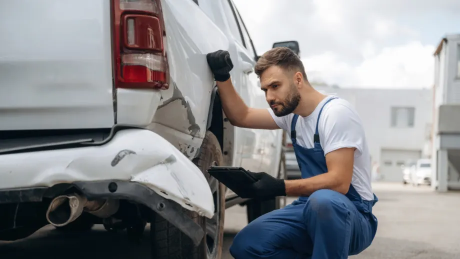 Technician kneels beside a white commercial van, examining damage to the rear bumper and body panel while holding a tablet.