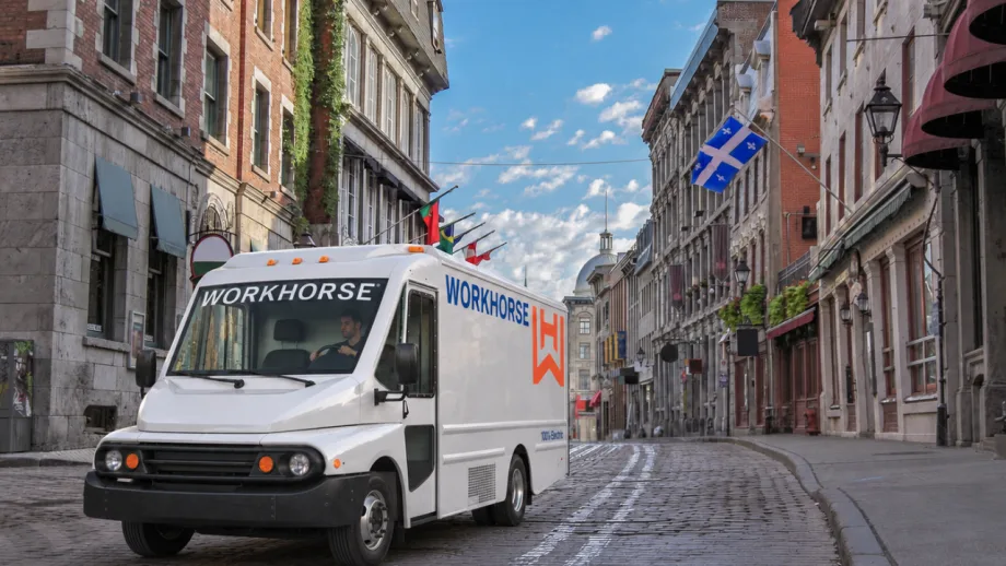 White Workhorse electric delivery truck driving along a cobblestone street lined with historic buildings and flags in a Canadian city. The truck features Workhorse branding and an orange logo on its side.