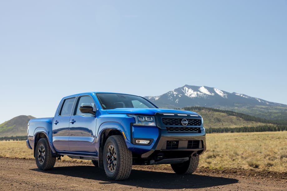 Blue 2027 Nissan Frontier Sport Edition pickup truck parked on a dirt road with snow-capped mountains in the background, showcasing its off-road styling and all-terrain tires.
