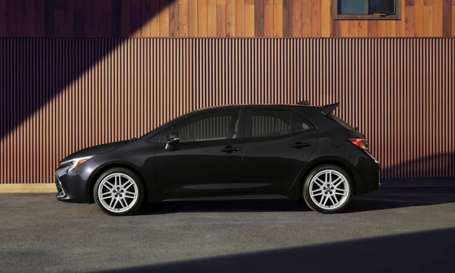 Side profile of a black 2027 Toyota Corolla Hatchback parked in front of a wood-paneled building, showing the compact hatchback's available 18-inch gloss white alloy wheels and updated styling.