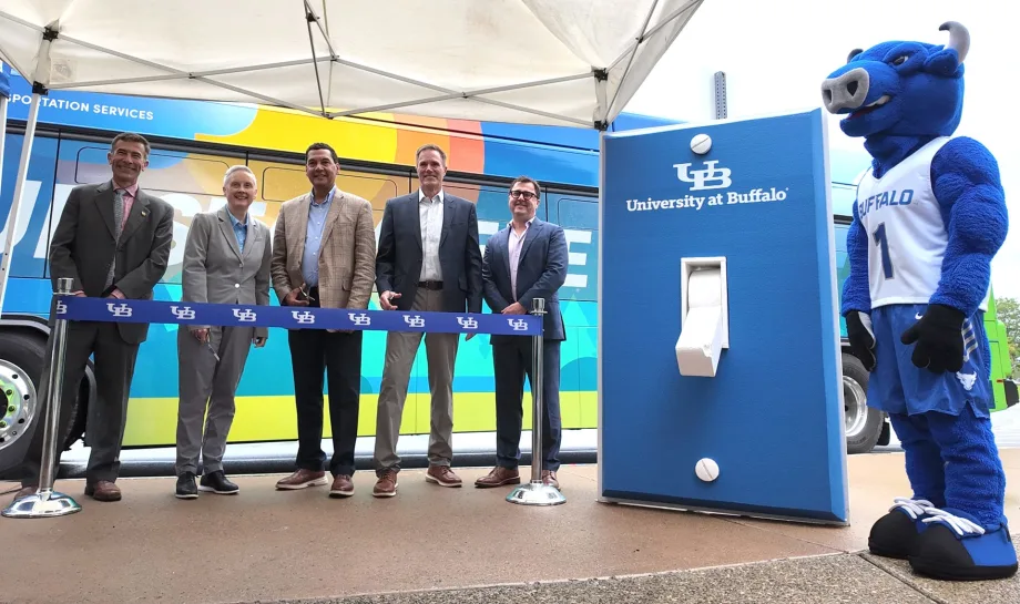 Five University at Buffalo representatives stand behind a ribbon beside a colorful transit bus and UB-branded charging station, with the Victor E. Bull mascot nearby.