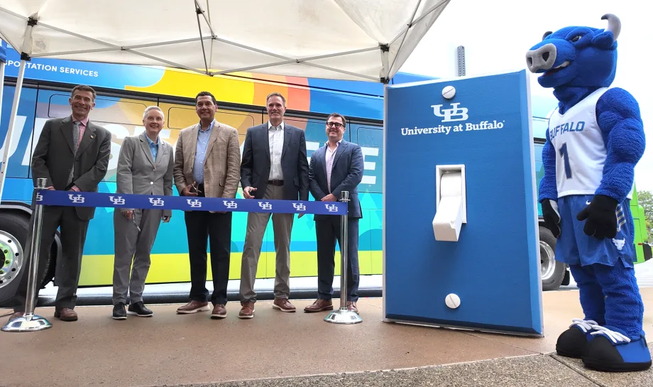 University at Buffalo officials stand beside a colorful transit bus and a UB-branded charging station during a ribbon-cutting event, with the university’s Victor E. Bull mascot nearby.