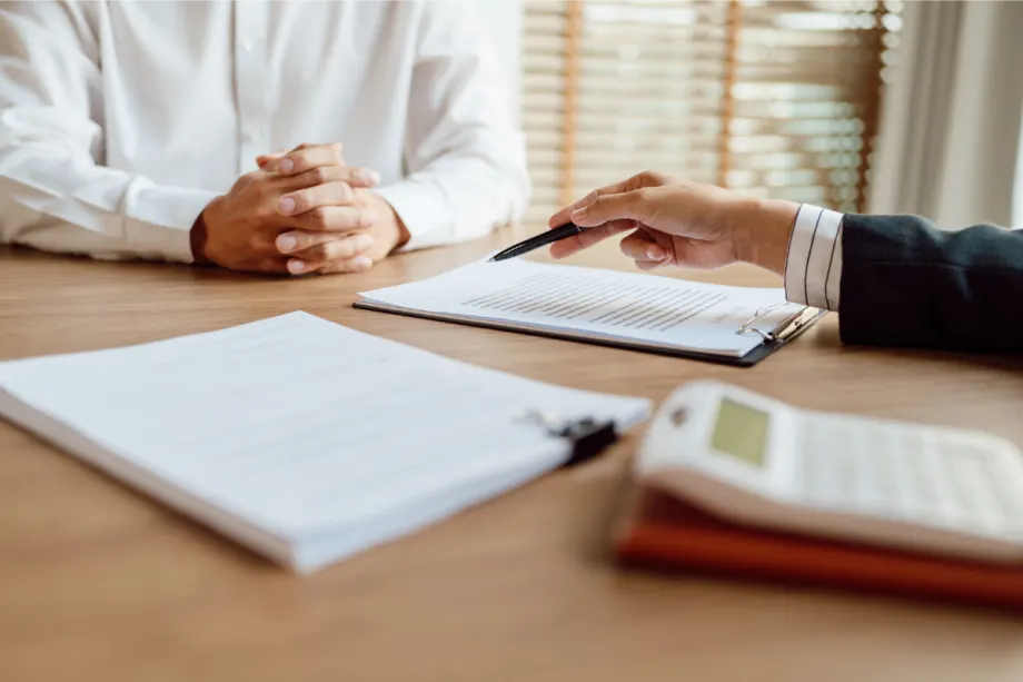 Two people sit across from each other at a desk during a business meeting. One person, wearing a white shirt, has their hands folded while the other gestures with a pen toward documents clipped to a clipboard. Additional paperwork and a calculator are visible on the table, suggesting a discussion involving contracts, finances, or administrative paperwork. Sunlight filters through window blinds in the background, creating a professional office setting.
