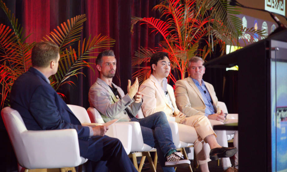 Moderator and three panelists on stage in white chairs in front of a red curtain and palm frond planters.
