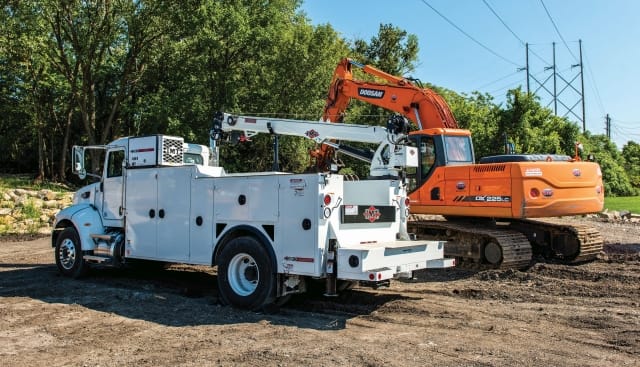White utility service truck with a crane parked beside an orange excavator at a roadside work site.