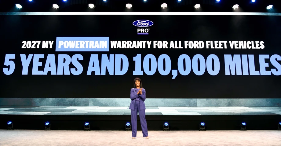 Presenter stands onstage before a large screen announcing Ford Pro’s expanded 5-year, 100,000-mile powertrain warranty for additional fleet vehicles beginning with the 2027 model year.