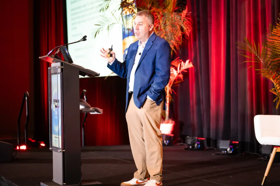 Speaker John Healy on stage with a podium, screen, and red curtains in the background.