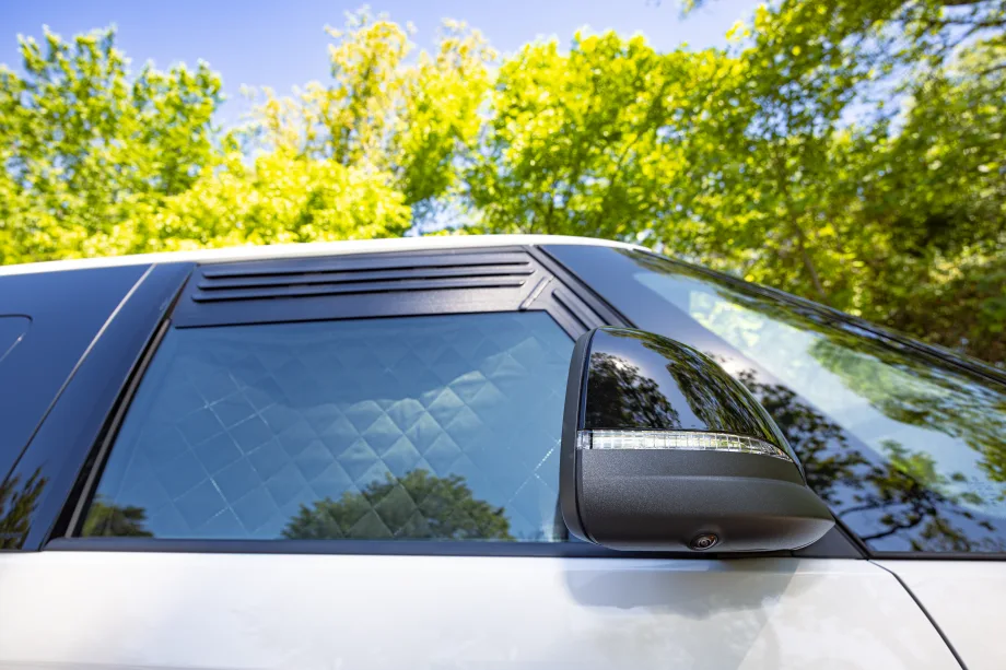Close-up of the 2027 Volkswagen ID. Buzz side window with quilted privacy shades and integrated side mirror camera parked outdoors.