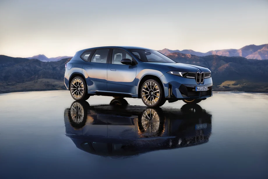 Blue BMW iX3 electric SUV parked on a reflective surface with mountain scenery in the background at dusk.