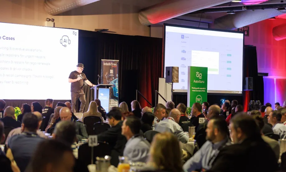 A presenter speaks on stage at a conference, addressing an audience seated at round tables, with large screens displaying presentation slides in the background.
