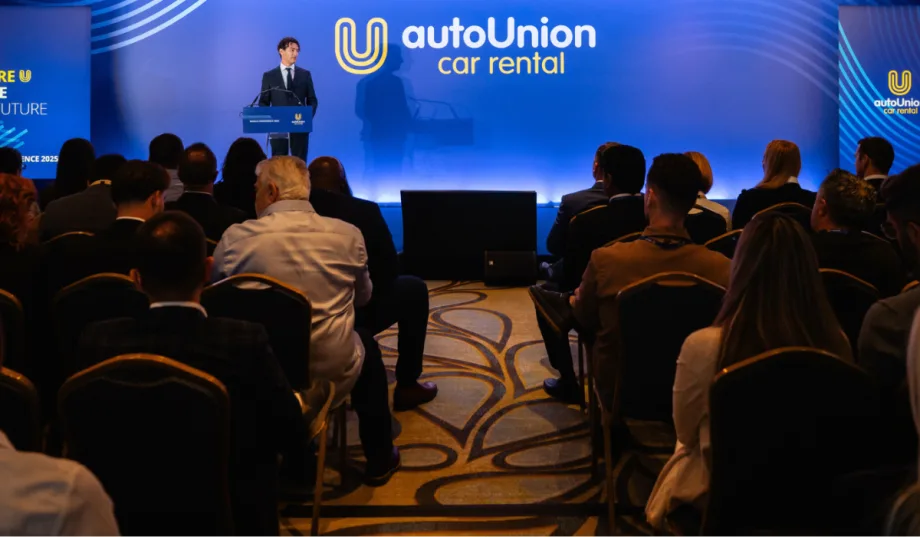 AutoUnion COO speaks to audience in front of wide blue screen with company logo on stage.