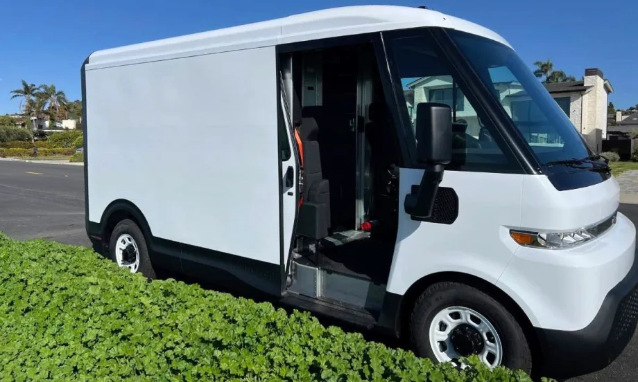 A white electric delivery van parked along a residential curb with low green shrubs.