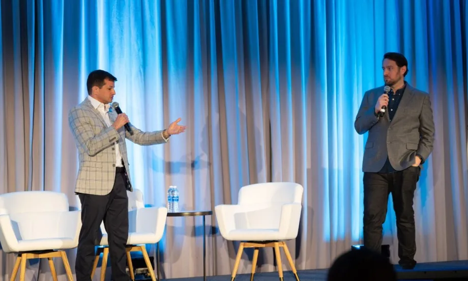 Two speakers standing on a stage talking at each other with a white chair between them.