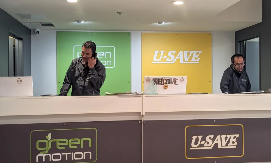 Two men working at a Green Motion U-Save car rental county with green and yellow logo backdrops.