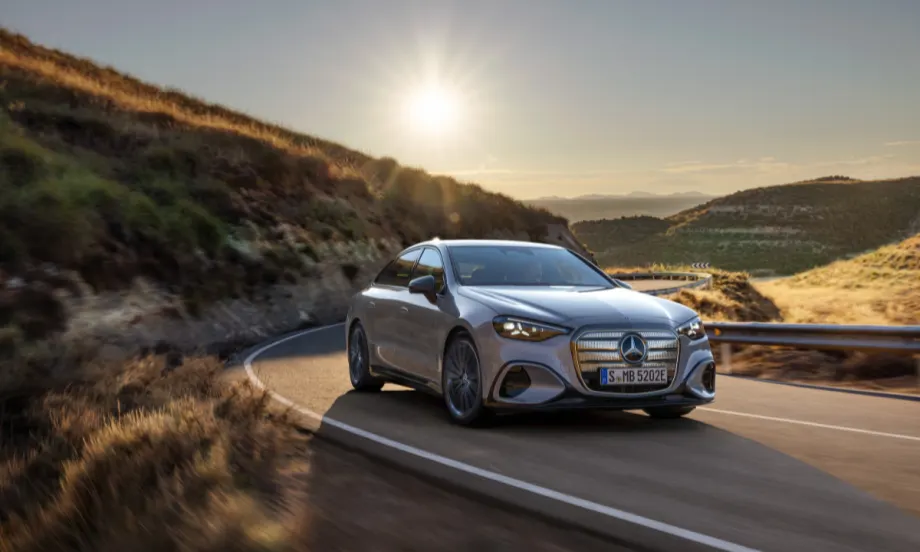 Front view of a silver Mercedes-Benz C-Class EV driving on a curving road with mountains and sunlight in the background.
