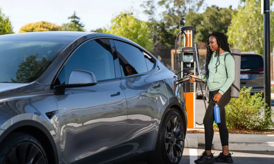 Person charging an electric vehicle at a public EV charging station, holding the charging connector next to a parked electric car outdoors.