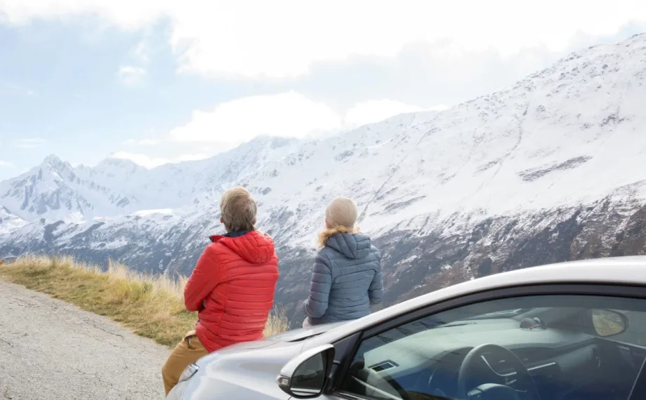 A couple sitting on the hood of a rental car looks at the scenic snowy mountain range.