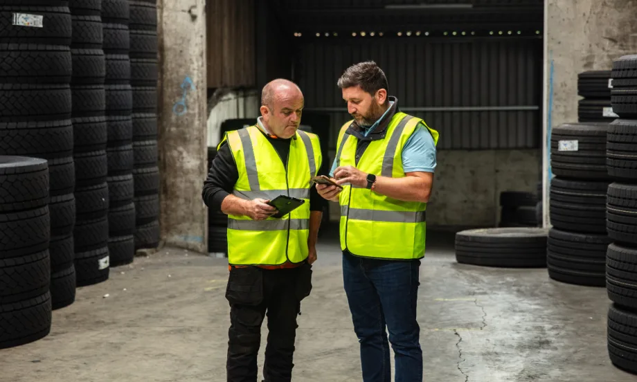 Two workers in safety vests look at a tablet and phone in a tire warehouse.