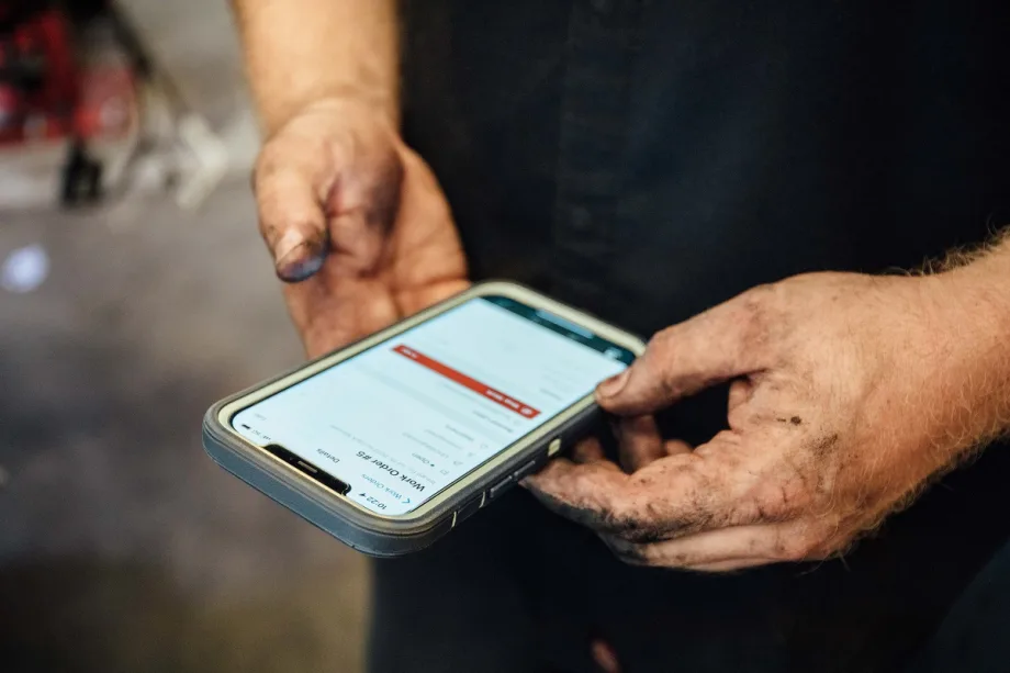 Grease-stained hands hold a smartphone displaying a digital fleet maintenance or vehicle service form.