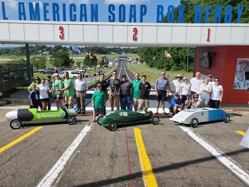 A group of participants and organizers stand behind three colorful soap box cars at the starting line of the All-American Soap Box Derby track in Akron, Ohio, celebrating the launch.