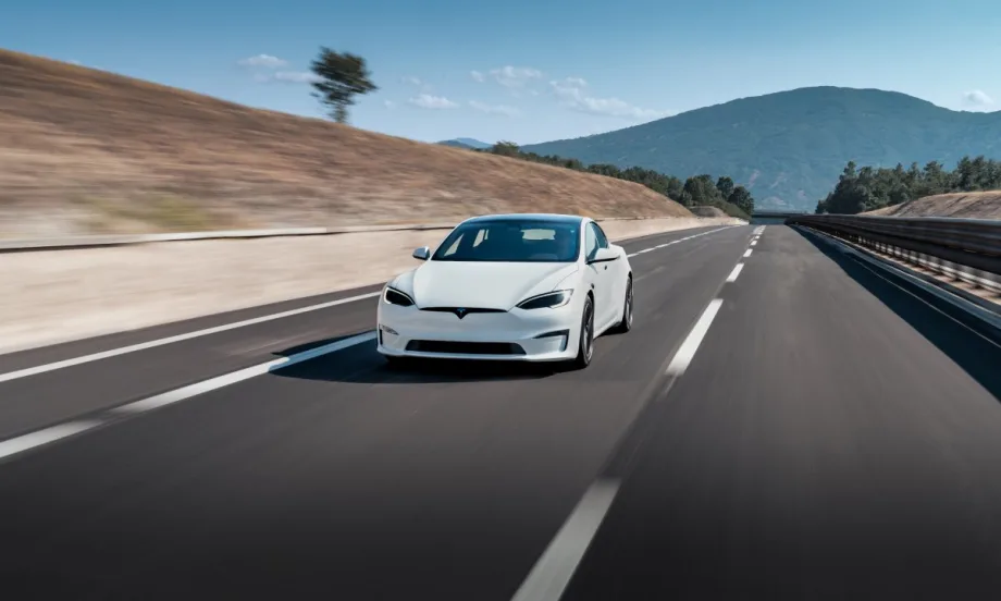 A white Tesla Model S driving down a paved road with hills and mountain in background on a sunny day.