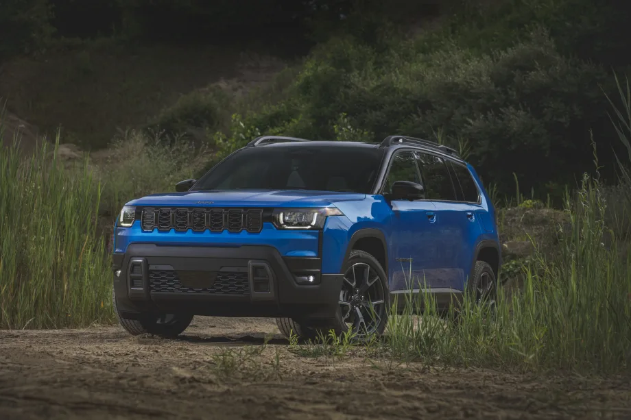 A blue Jeep Cherokee SUV parked outdoors on a dirt trail is surrounded by tall grass and greenery.