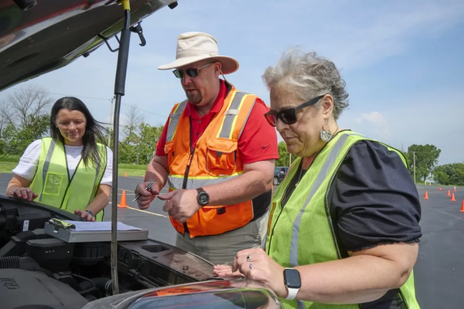 A man in an orange vest shows two women in green vests items under the hood of a vehicle.