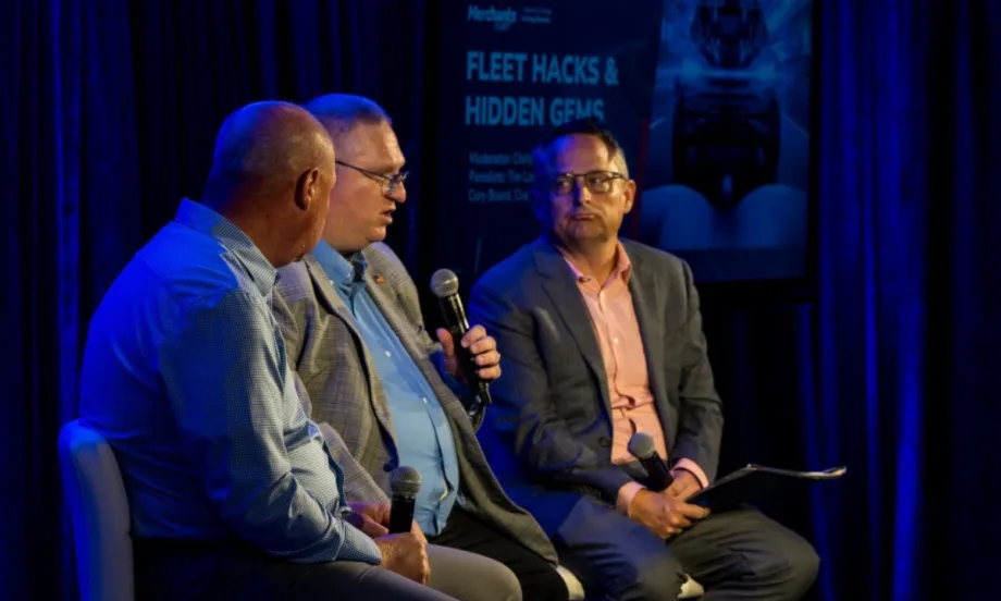 Cory Beard holds a mic next to Tim Lovett and Chris Brown, who sit on a stage for a panel discussion at the Merchant's Fleet conference in New Hampshire.