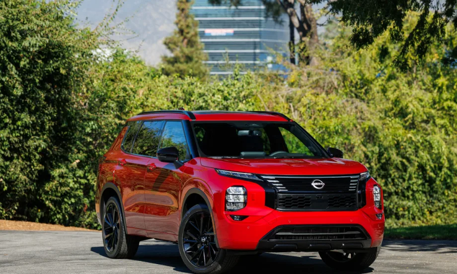 A red 2026 Nissan Rogue plug-in hybrid drives on a road next to bushes.