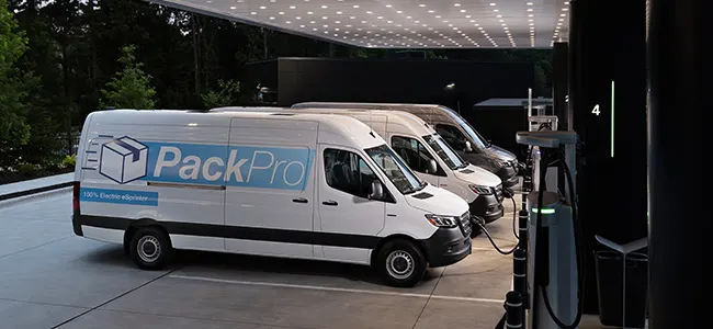 Row of white electric delivery vans labeled ‘PackPro’ charging at a modern outdoor EV station .