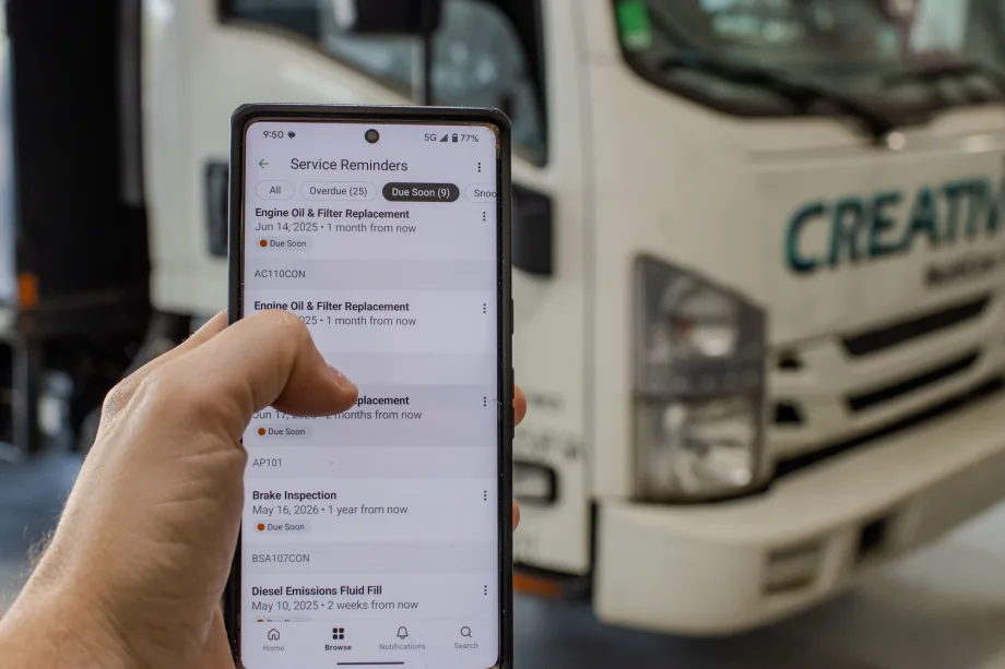 A person holds a smartphone displaying a list of vehicle service reminders, including engine oil and filter replacement, while standing in front of a white commercial truck.