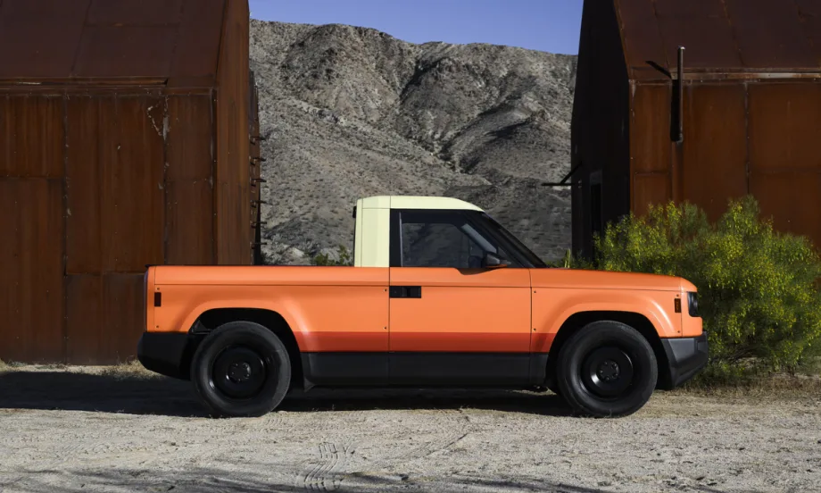 Orange Slate electric pickup truck parked outdoors between industrial structures, representing the company’s upcoming customizable EV supported by new funding.