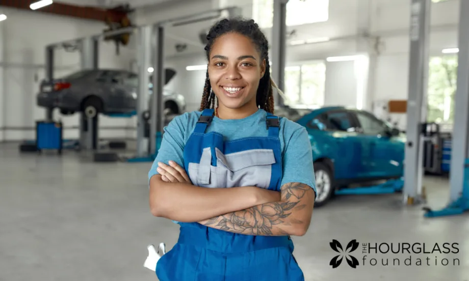 A woman in a blue shirt and apron stands in an automotive suite with her arms crossed.